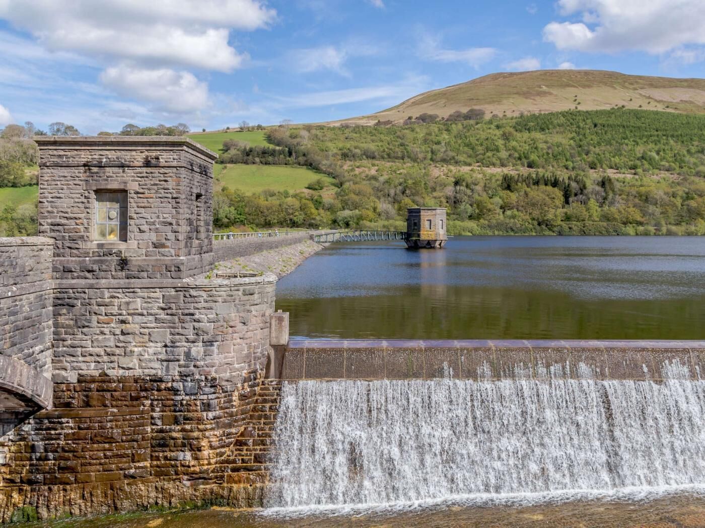 Ein steinerner Damm mit einem kleinen Turm, einem Wasserfall und einem See mit Hügeln im Hintergrund.