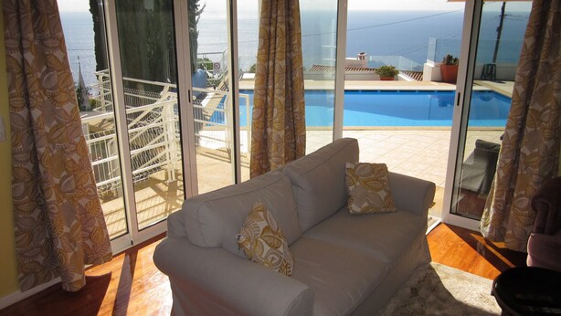 Ein Wohnzimmer mit einer Couch, Blick auf einen Pool und ein Balkon mit Geländer.