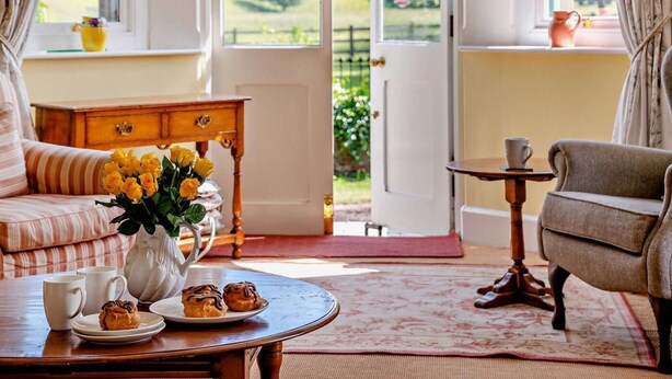 A cozy room with a sofa, a table with pastries, a vase with flowers, and a view of greenery through a door. - 3 Bed in Brecon (oc-bn134) (Brecon)