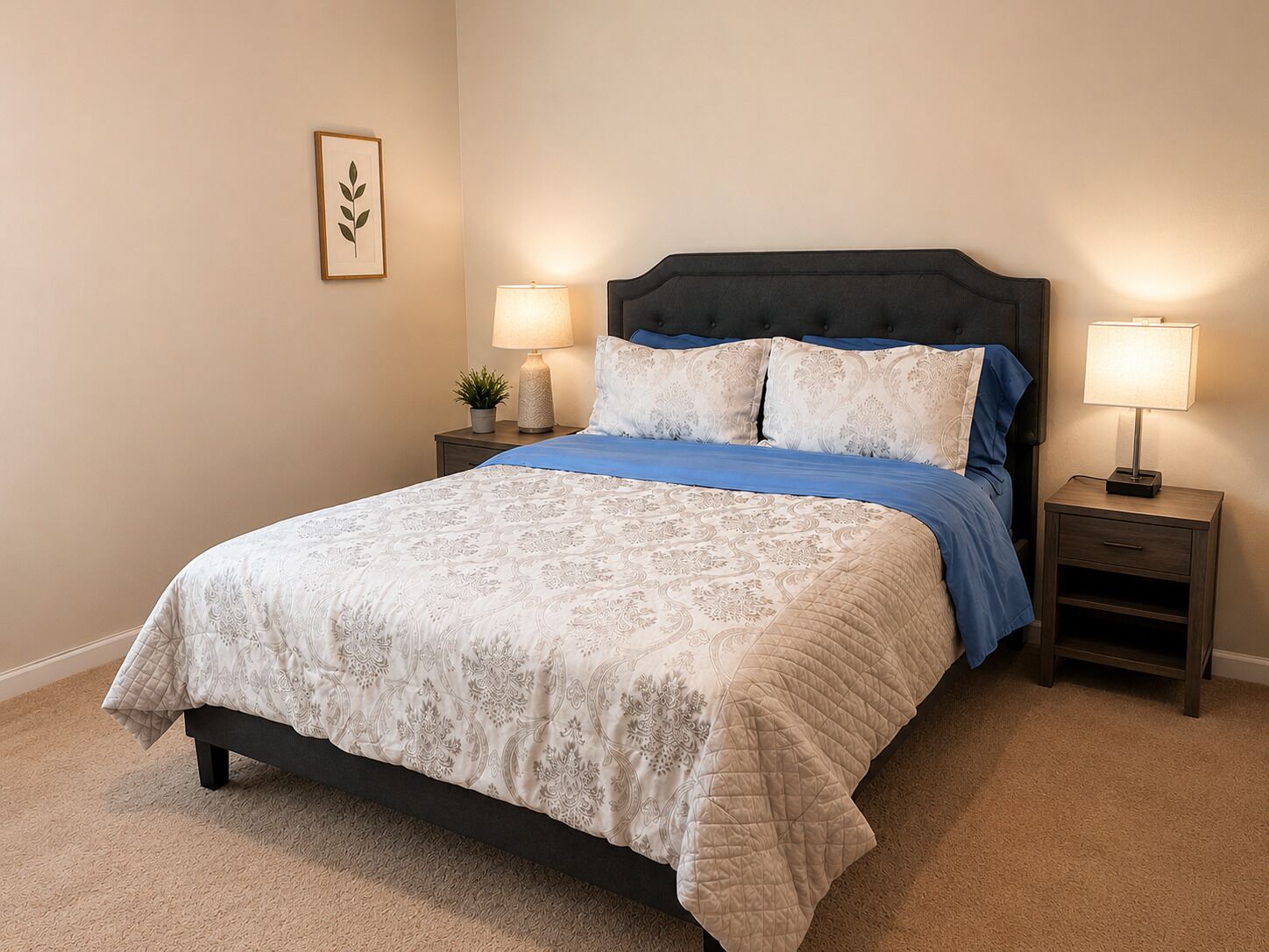 A neatly made bed with a patterned bedspread, two bedside lamps, a small potted plant, and a framed picture on the wall.