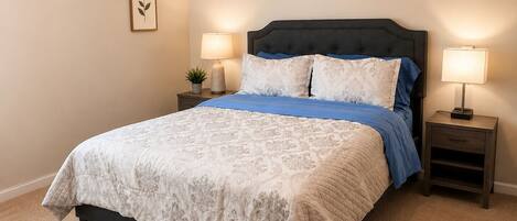 A neatly made bed with a patterned bedspread, two bedside lamps, a small potted plant, and a framed picture on the wall.