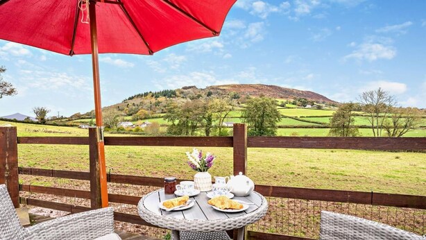 Una mesa de mimbre con platos de comida, un paraguas rojo y una vista panorámica de un paisaje verde con colinas.
