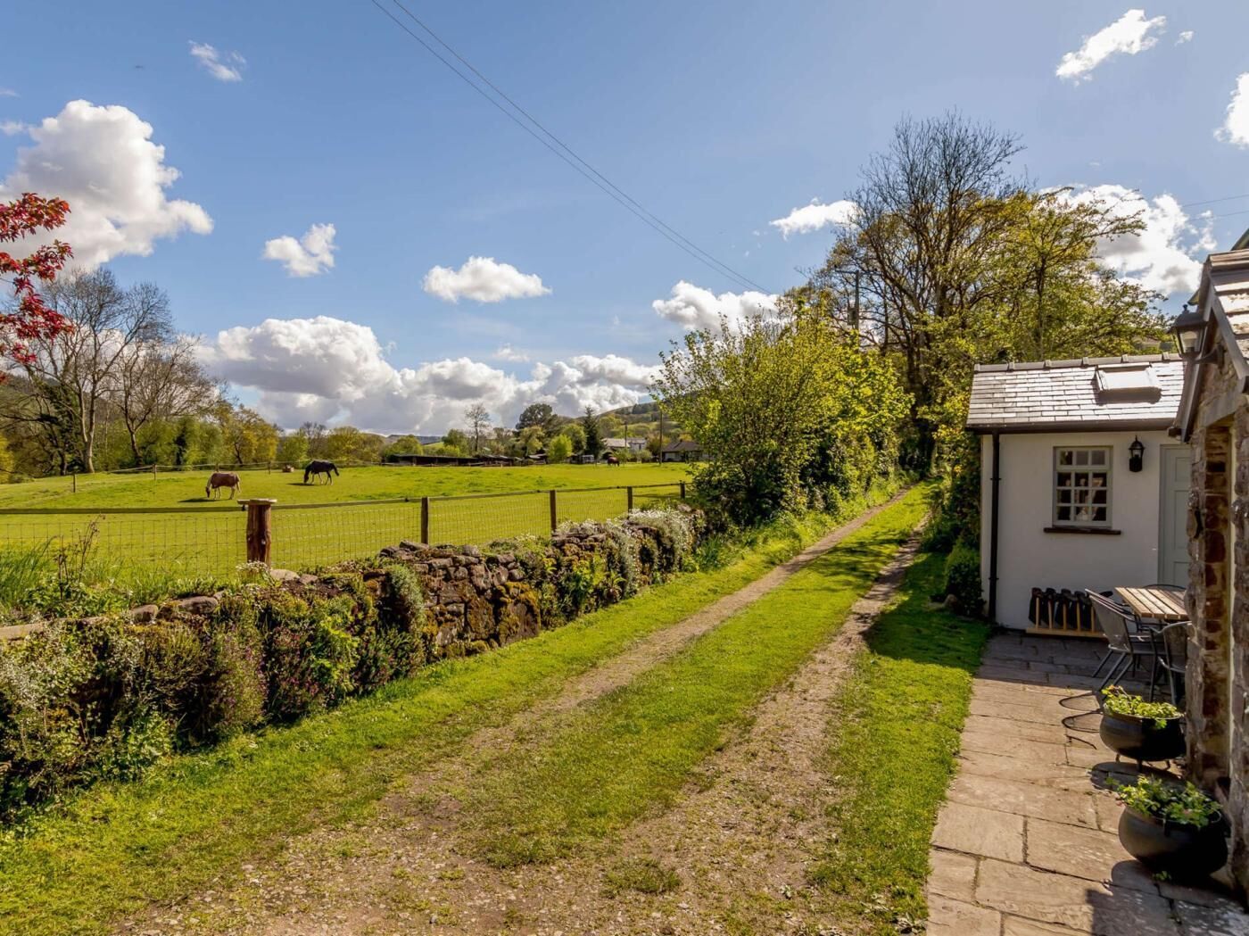 A rural property with a stone pathway, a fenced field with grazing animals, and a small white building with outdoor seating.