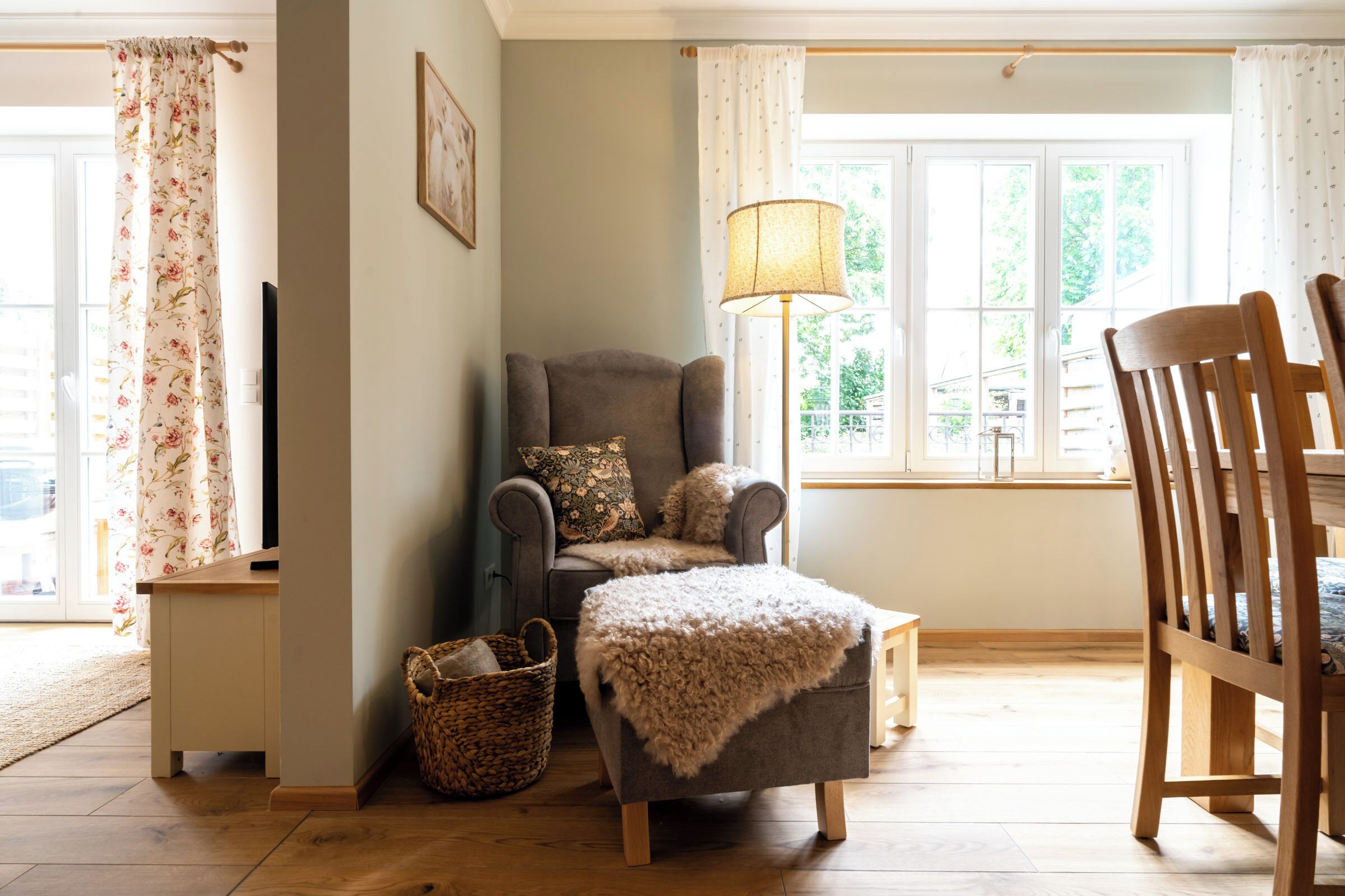 A cozy living room with a grey armchair, a wooden chair, a floor lamp, and a window with curtains.
