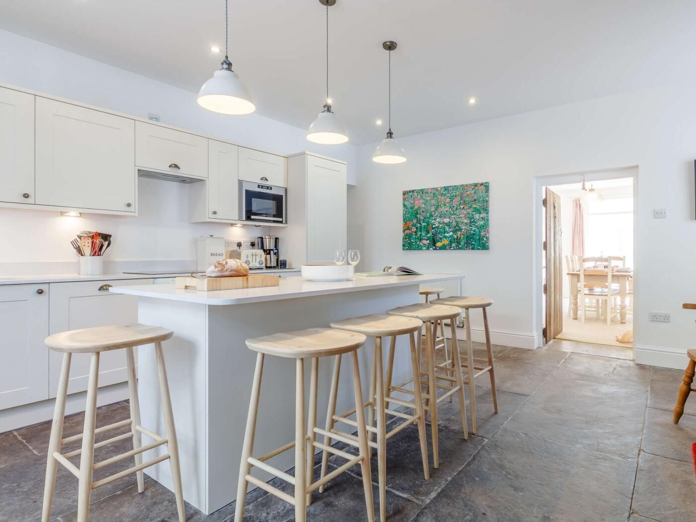 A modern kitchen with white cabinets, a central island with stools, pendant lighting, and a dining area visible through an open door.