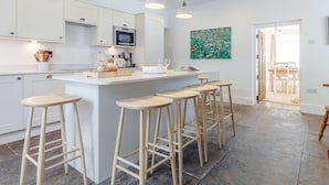 Een moderne keuken met witte kasten, een centraal eiland met krukken, hangende lampen en een zichtbare eethoek door een open deur.