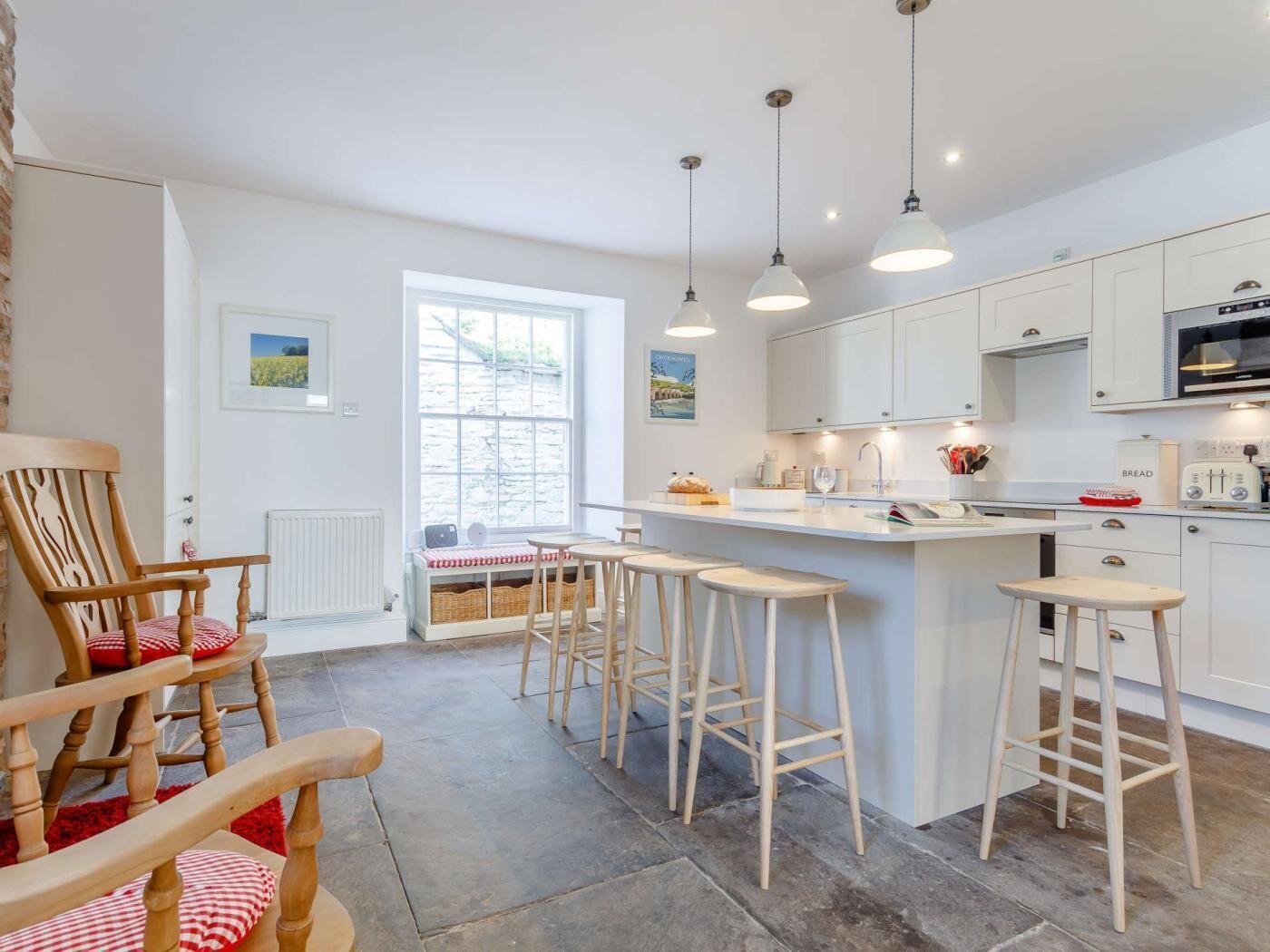 A modern kitchen with white cabinets, a central island with stools, pendant lighting, and a view through a large window.
