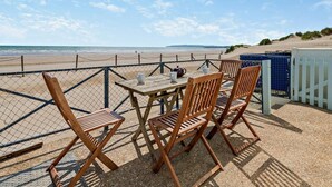 Une table et des chaises en bois sont disposées sur une terrasse en gravier, offrant une vue sur la plage et l’océan.