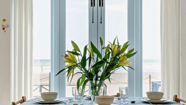 Une table à manger recouverte d’une nappe à carreaux, un vase rempli de fleurs jaunes et une vue sur la plage, visible à travers de grandes fenêtres.