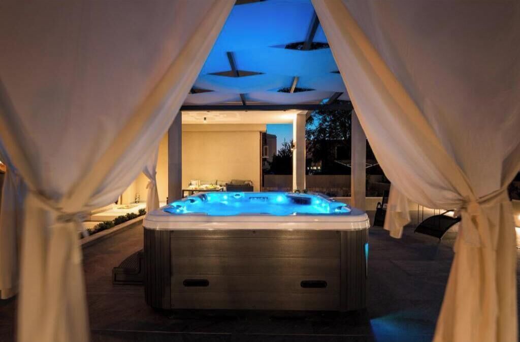 Un jacuzzi avec un éclairage bleu, entouré de rideaux blancs, et une vue sur un bâtiment en arrière-plan.