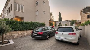Ein Parkplatz mit zwei Autos, ein Gebäude mit Natursteinfassade und einer Hecke.