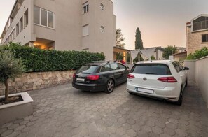 Ein Parkplatz mit zwei Autos, ein Gebäude mit Natursteinfassade und einer Hecke.