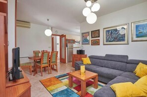 A living room with a gray sofa, wooden coffee table, and a colorful rug. There is a dining area with wooden chairs and a table. The room has artwork on the walls and a ceiling light fixture.