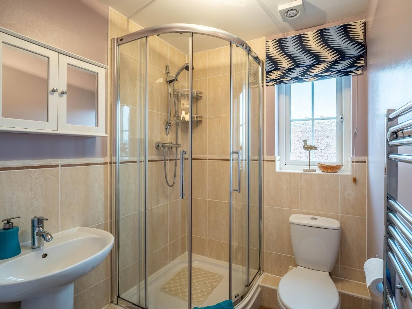 A bathroom with a glass-enclosed shower, a toilet, a sink, and a window with a leaf-patterned curtain.