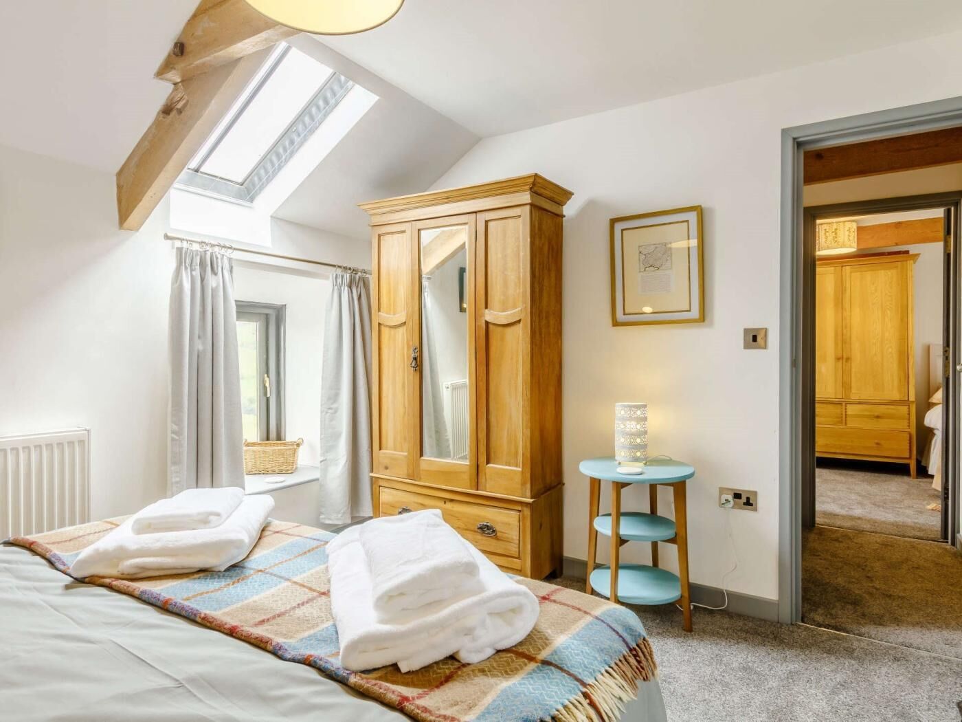 Un dormitorio con cama, armario de madera, mesita con lámpara, ventana con cortinas y puerta que da a otra habitación.