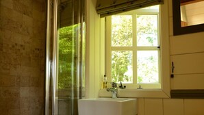 Ein Badezimmer mit Duschkabine aus Glas, einem Fenster mit Blick auf Bäume und einem weißen Waschbecken mit Armatur.