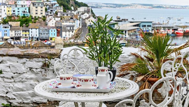 Eine Terrassennische am Meer mit gedecktem Tisch für Tee, mit Blick auf eine bunte Küstenstadt und das Meer.