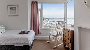 Ein Schlafzimmer mit Bett, Sessel, Kommode, einem Fenster mit Blick auf eine Stadtansicht und einer Deckenleuchte.
