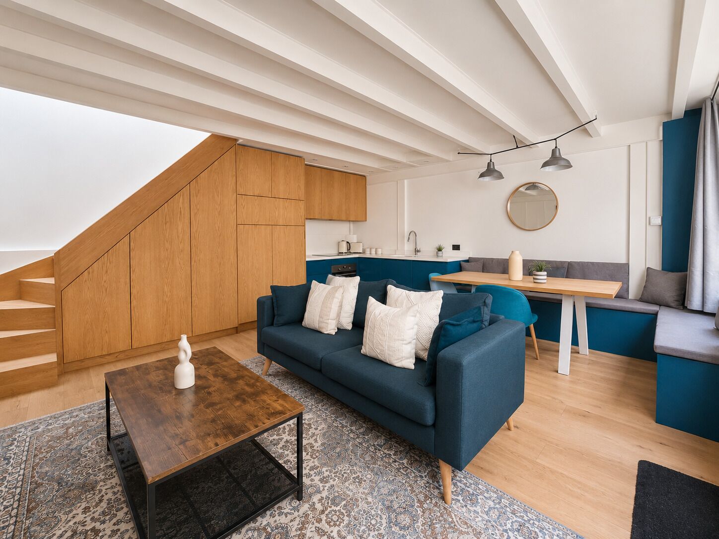 A modern living space with a wooden staircase, a blue sofa set, a wooden coffee table, and a kitchen area with a sink and a round mirror.
