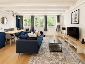 Een moderne woonkamer met een bank, salontafel en televisie. Er is een eethoek met een tafel en stoelen, en een groot raam met gordijnen.