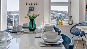 Un coin repas avec une table dressée pour quatre personnes, un vase vert orné de fleurs et une vue sur la ville à travers de grandes fenêtres.