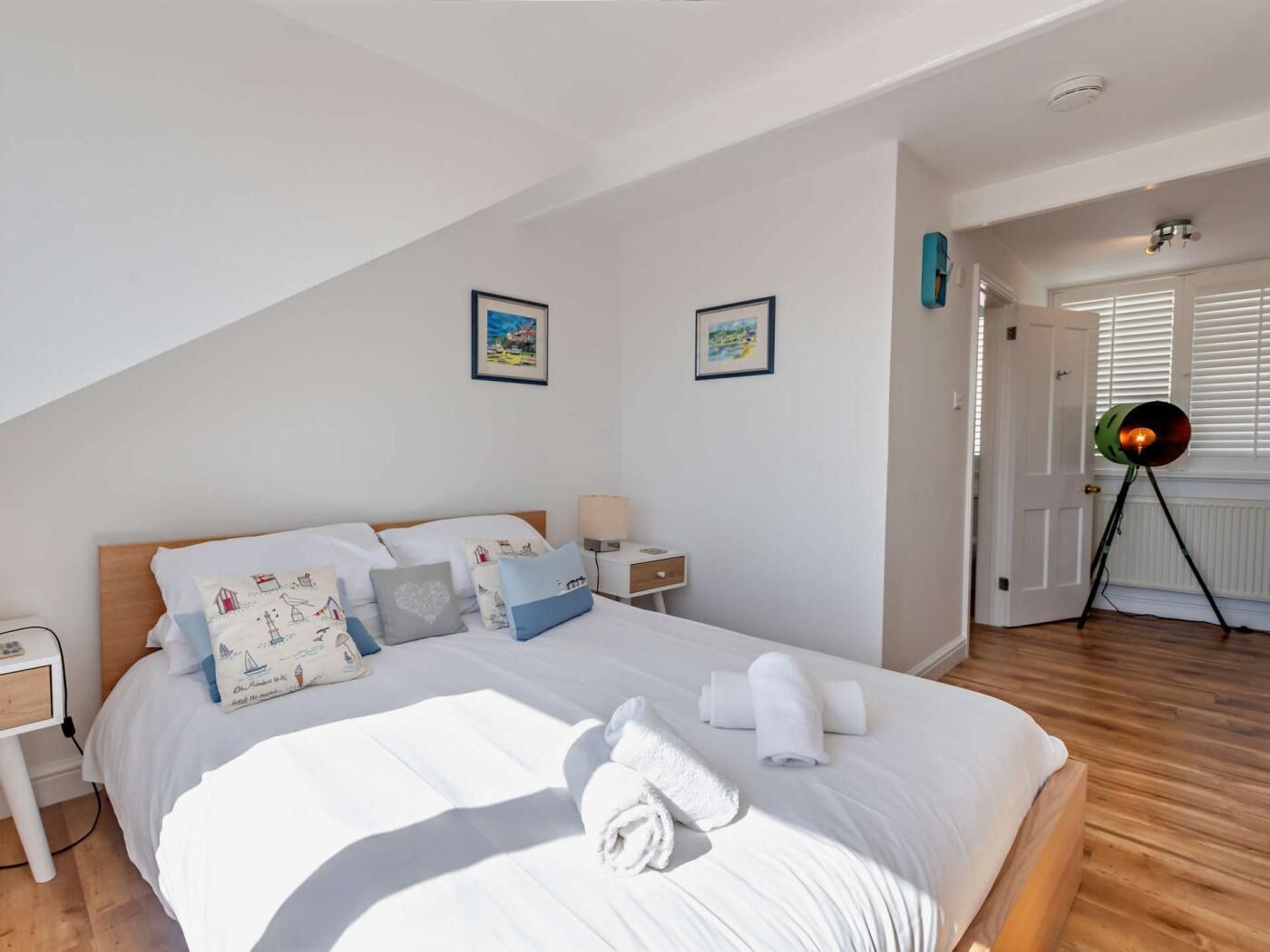 Uma cama bem arrumada com lençóis brancos e almofadas decorativas, uma mesa de cabeceira com abajur, e um quadro na parede.