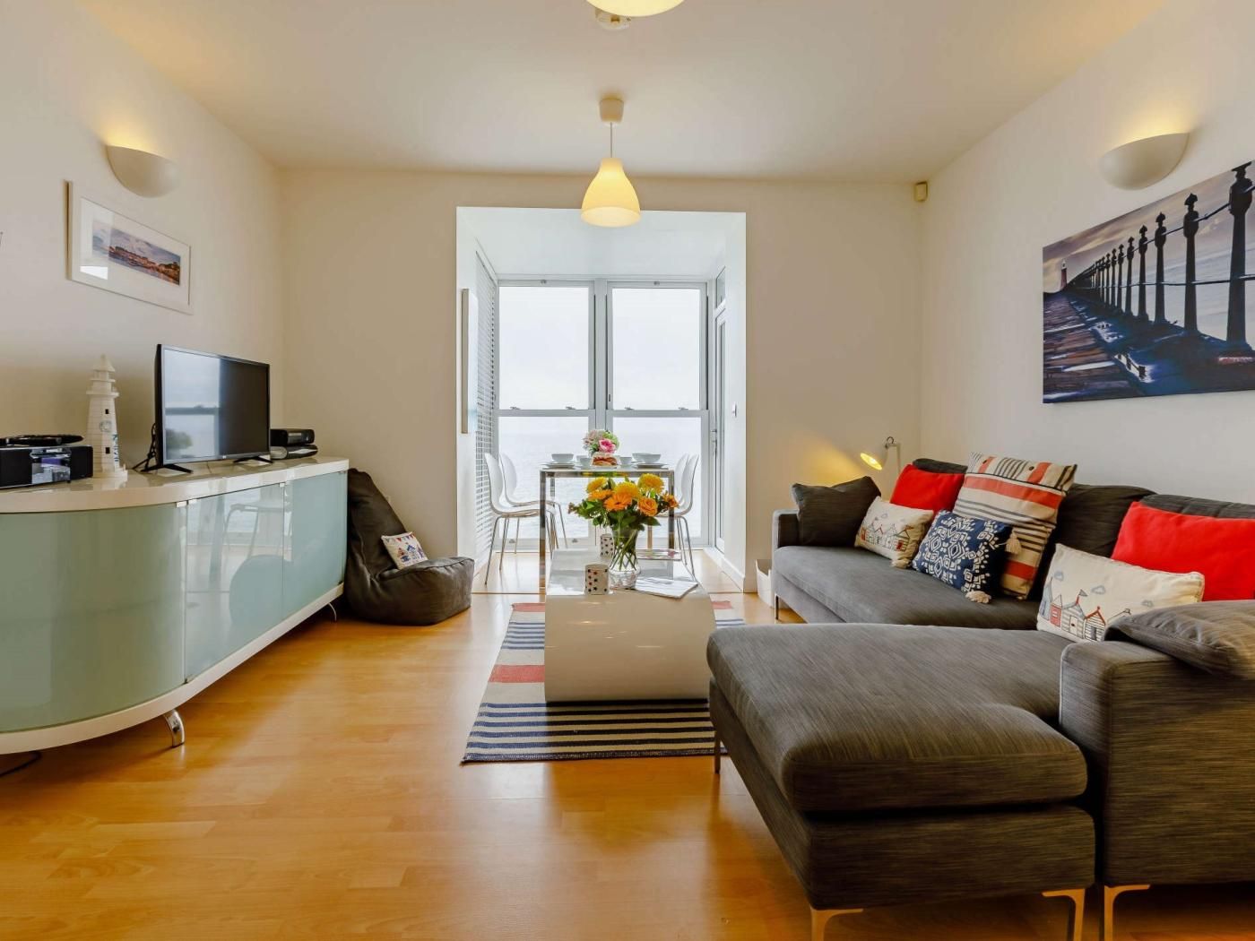 A modern living room with a sofa, a coffee table, and a TV. There is a dining area with a table and chairs. The room has a large window and a view of the outside.