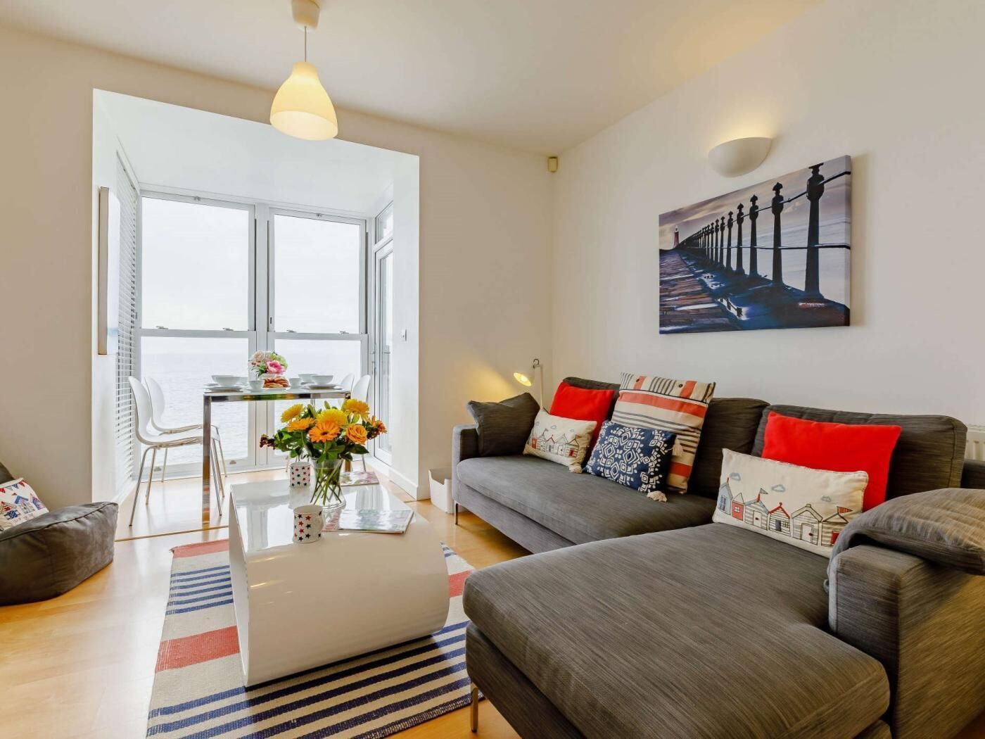 A modern living room with a sofa, a dining table with chairs, a vase of flowers, and a painting on the wall.