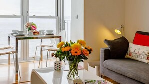 A room with a dining table, chairs, a sofa, a lamp, and a view through large windows. - 2 Bed in Ilfracombe (oc-caps8) (Ilfracombe)