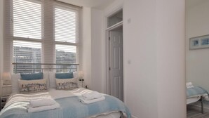 A bedroom with a bed, two bedside tables, a window with blinds, a door, and a carpeted floor. - 2 Bed in Ilfracombe (oc-caps8) (Ilfracombe)