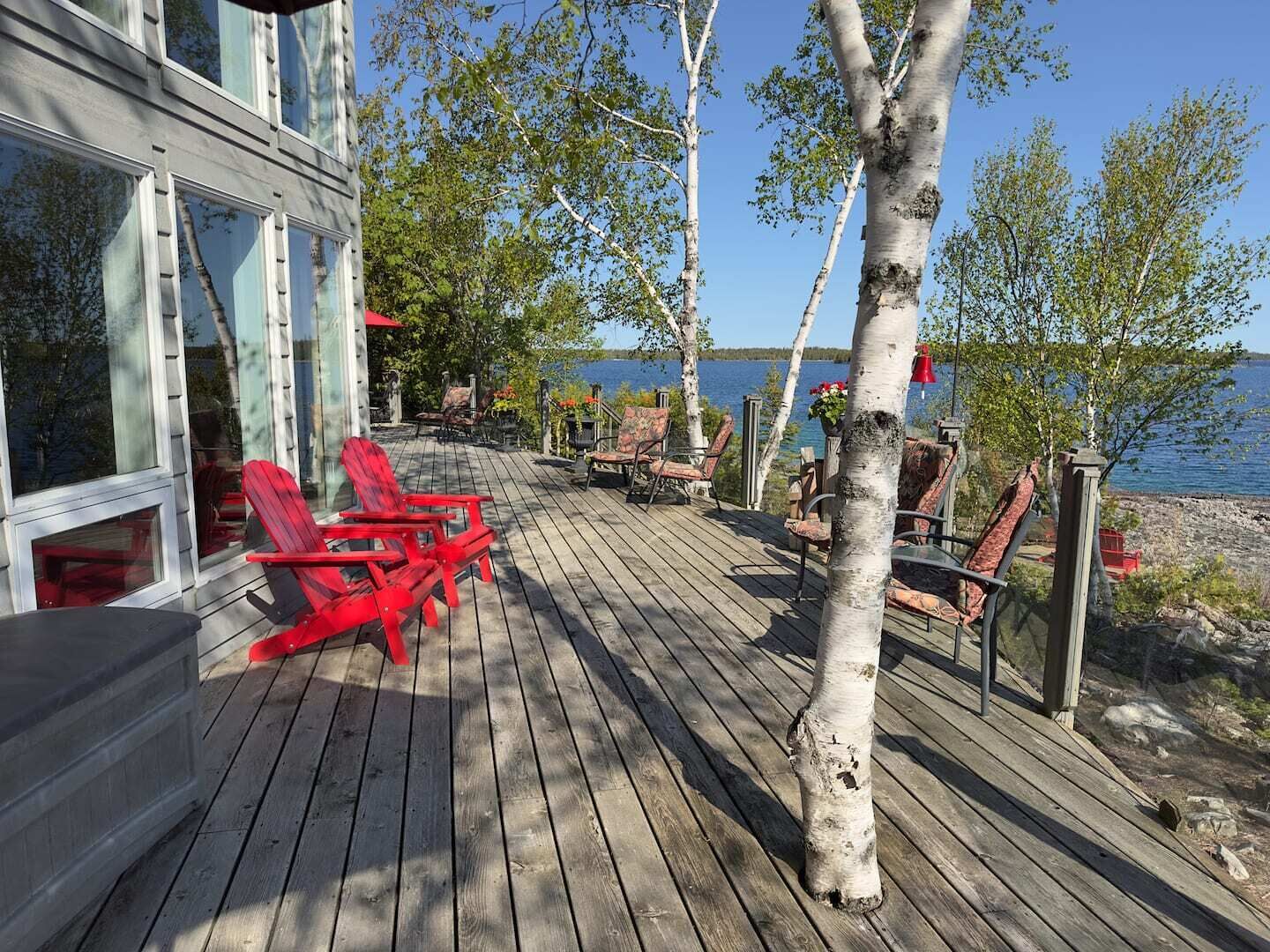 Terraza de madera con sillas Adirondack rojas, vista a un lago y árboles de abedul.