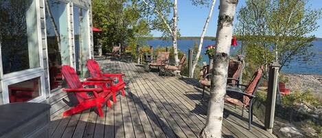 Terraza de madera con sillas Adirondack rojas, vista a un lago y árboles de abedul.