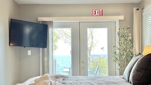 Una camera da letto con un letto, una TV fissata al muro, un cartello di uscita, vista mare attraverso una porta e una pianta.