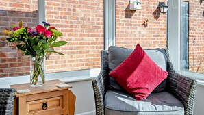 Een kamer met een bakstenen muur, een rieten stoel met een rood kussen, een houten nachtkastje met een vaas bloemen, en een raam met een gestreept gordijn.