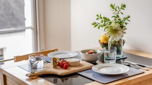 Uma mesa de jantar com um prato, uma tigela, um copo de água, uma tábua de corte de madeira com queijo e tomates, e um vaso com flores.