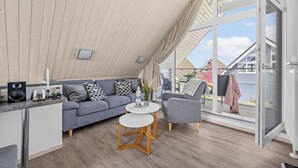 Et moderne loftrum med skråvæg, en grå sofa, et rundt hvidt bord og et stort vindue med udsigt udad.