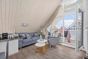 Ein stilvolles Loft-Zimmer mit schrägem Dach, einem grauen Sofa, einem runden weißen Tisch und einem großen Fenster mit Blick ins Freie.