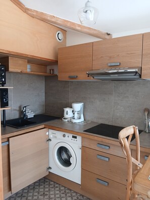 A compact kitchen with wooden cabinets, a built-in washing machine, a coffee maker, and a wooden chair. - Charming studio apartment in the Cassy harbor (Lanton)