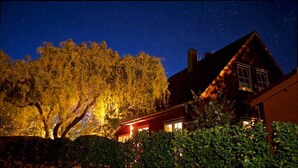 Una casa con un muro de piedra al frente, un árbol grande a la izquierda y un cielo nocturno lleno de estrellas.