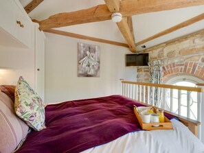 A bedroom with a stone fireplace, a bed with a purple blanket, a wooden ceiling, and a TV mounted on the wall. - 1 Bed in Bedale (oc-g0044) (Patrick Brompton)