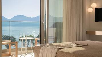A bedroom with a large bed, a glass door leading to a balcony with a view of a lake and mountains, and a small table with chairs.