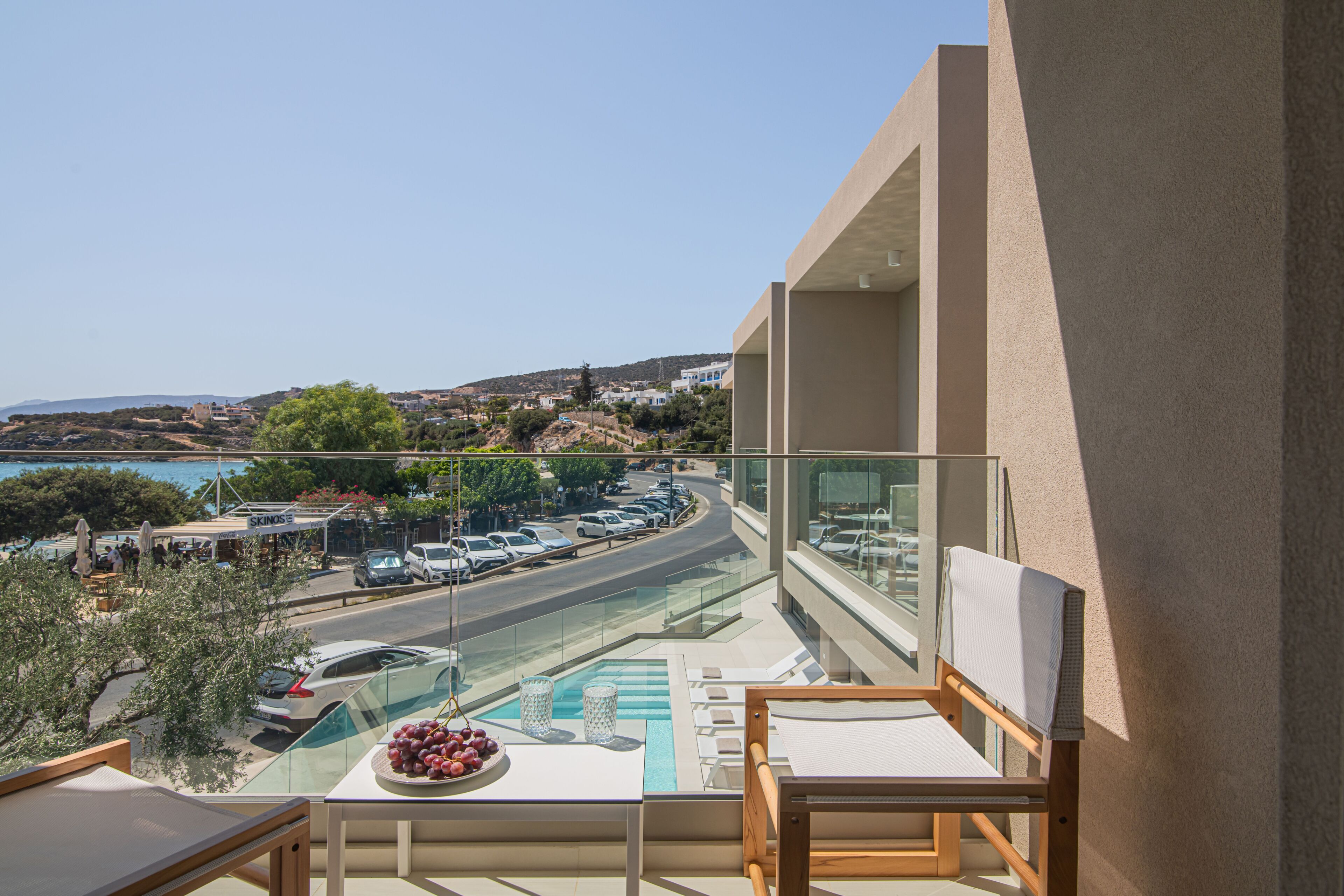 A balcony with a view of a coastal road, a swimming pool, and a dining area with a table and chairs.