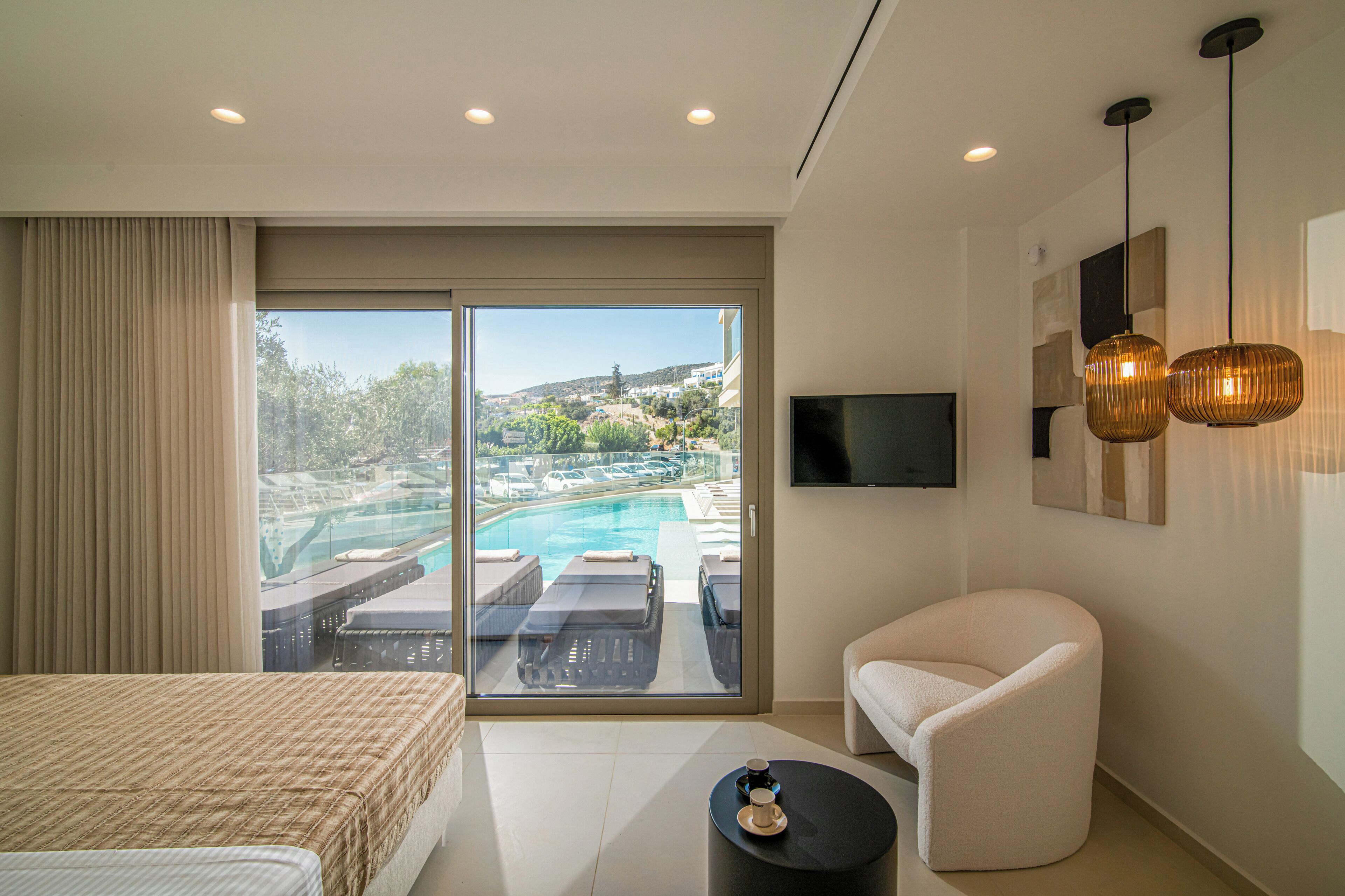 A modern hotel room with a large sliding glass door leading to a pool area, a flat-screen TV mounted on the wall, and a comfortable seating area with a sofa and a small round table.