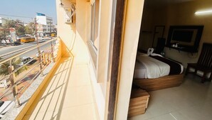 A balcony with a view of a busy street, a bed with a wooden frame, a television, and a ceiling fan. - Hotel Nageshwar Palace Rajgir (Rajgir)