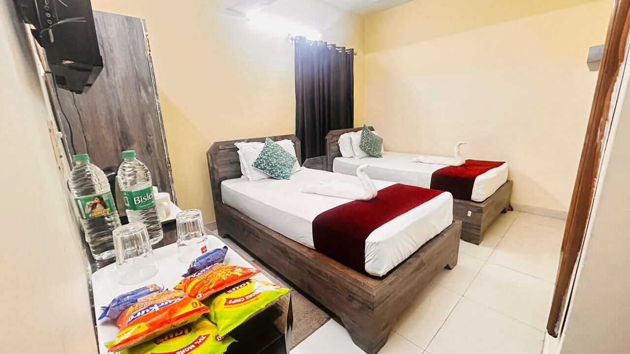 Habitación de hotel con tres camas, cabecera de madera, un espejo, una mesita con aperitivos y una botella de agua Bisleri.