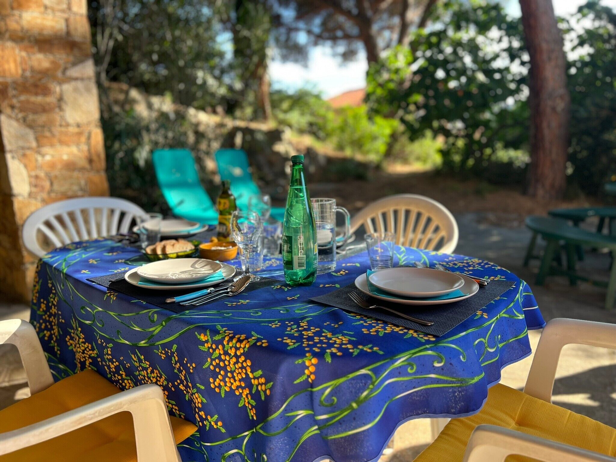 Mesa posta ao ar livre com toalha azul, pratos, talheres e uma garrafa verde.