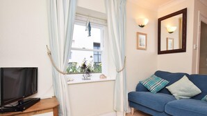 Ein Wohnzimmer mit Fernseher auf einem Holzboden, ein blaues Sofa mit Kissen, ein Fenster mit Vorhängen und eine kleine Pflanze im Fensterbrett.