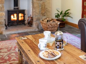 Ein Holztisch mit einer Auswahl an Gebäck, einer Kaffeekanne und einer Tasse in einem gemütlichen Raum mit Kamin und Pflanze.