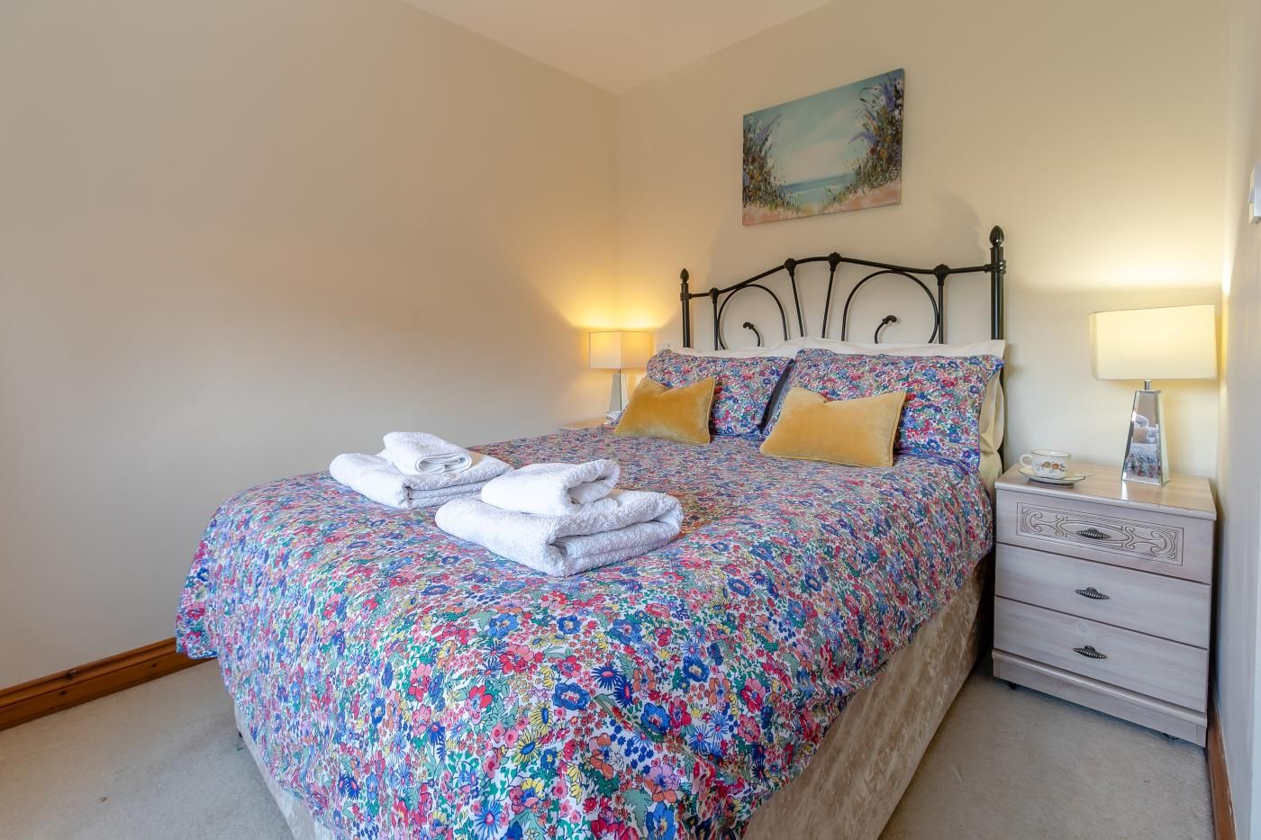 Un letto rifatto con cura, lenzuola e cuscini a motivi floreali, asciugamani piegati. Si nota un comodino con una lampada e un piccolo oggetto decorativo.
