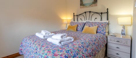 Un letto rifatto con cura, lenzuola e cuscini a motivi floreali, asciugamani piegati. Si nota un comodino con una lampada e un piccolo oggetto decorativo.